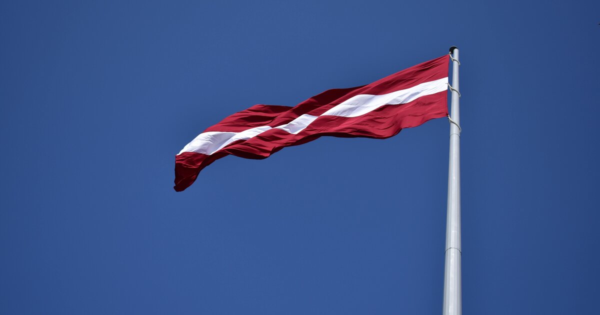 Le drapeau letton — rouge carmin et bande blanche — flottant sur ciel bleu.
